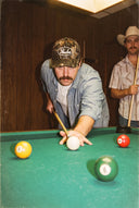 Man wearing the Paulie's Camo Cap lining up a pool shot in a dive bar